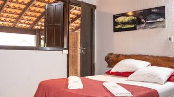 Habitación de hotel con cama, colcha roja, almohadas blancas, cabecera de madera, un cuadro en la pared y una ventana con techo de madera.