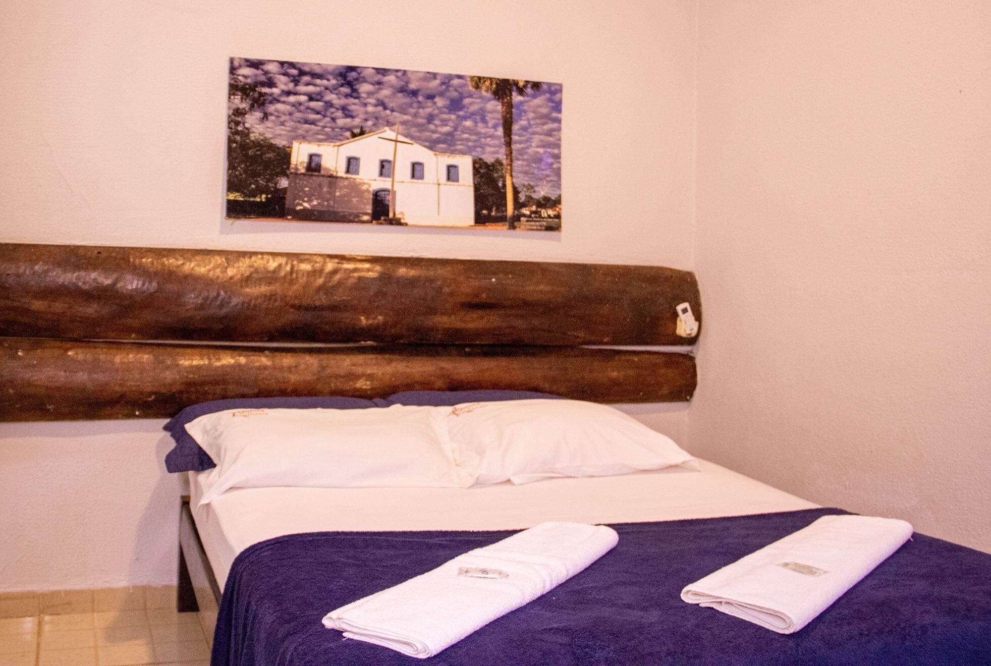 Quarto de hotel com cabeceira de madeira, cama com roupa de cama branca e manta roxa. Há um quadro de um edifício na parede.