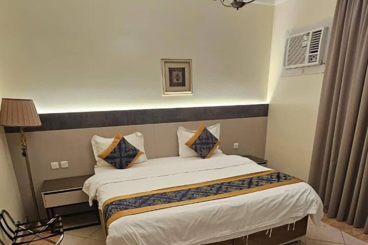 Une chambre d’hôtel avec un lit, un bureau, une chaise, une lampe, un tableau au mur et un luminaire au plafond.