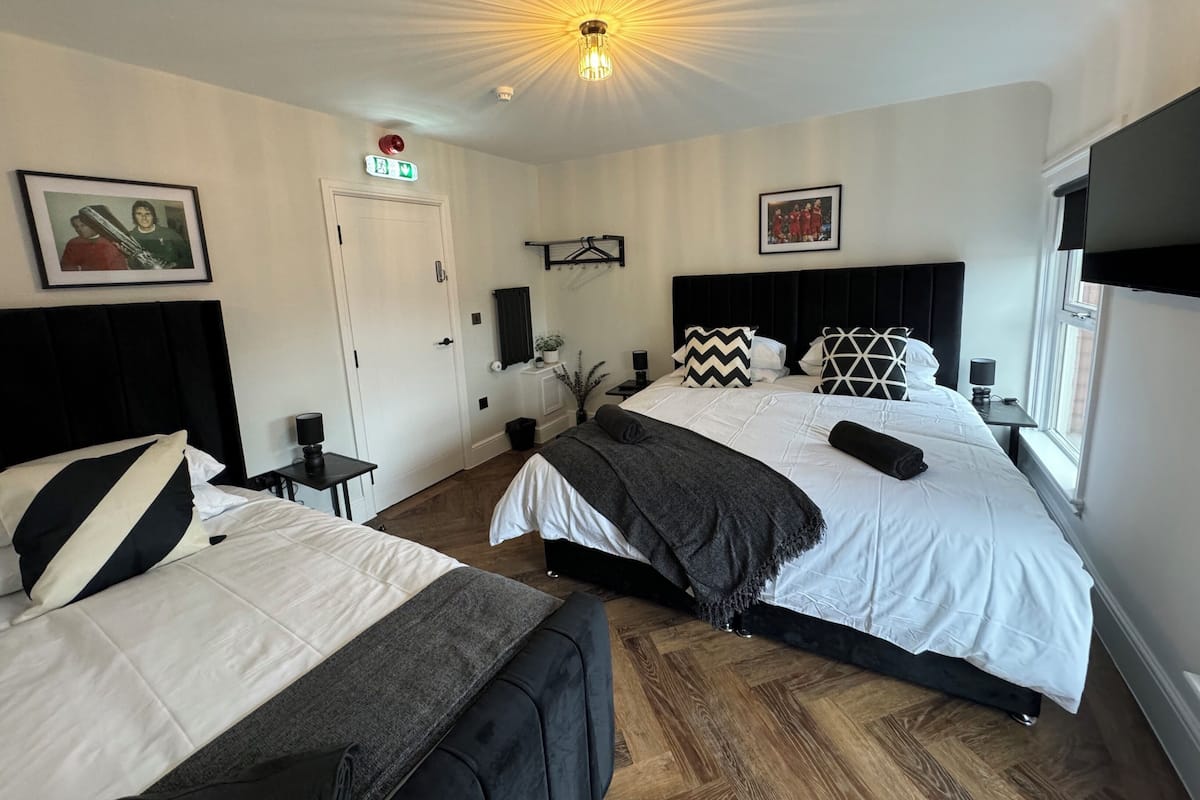 A hotel room with two beds, a ceiling light, a door, a small table with a lamp, and a window.