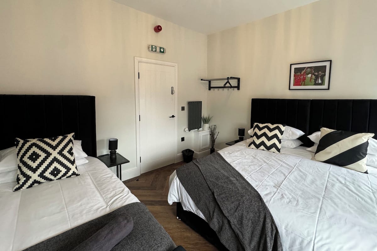 A hotel room with two beds, a door, a small table with a lamp, a wall-mounted shelf, and a framed picture.