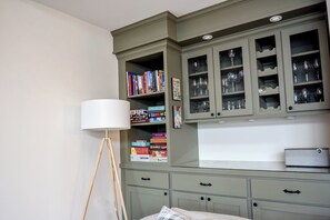 A room with a green cabinet containing books and glassware, a floor lamp, and a bed with a white headboard.