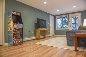 A living room with a retro arcade machine, a television on a wooden stand, a grey couch, and a large window with a view of trees.