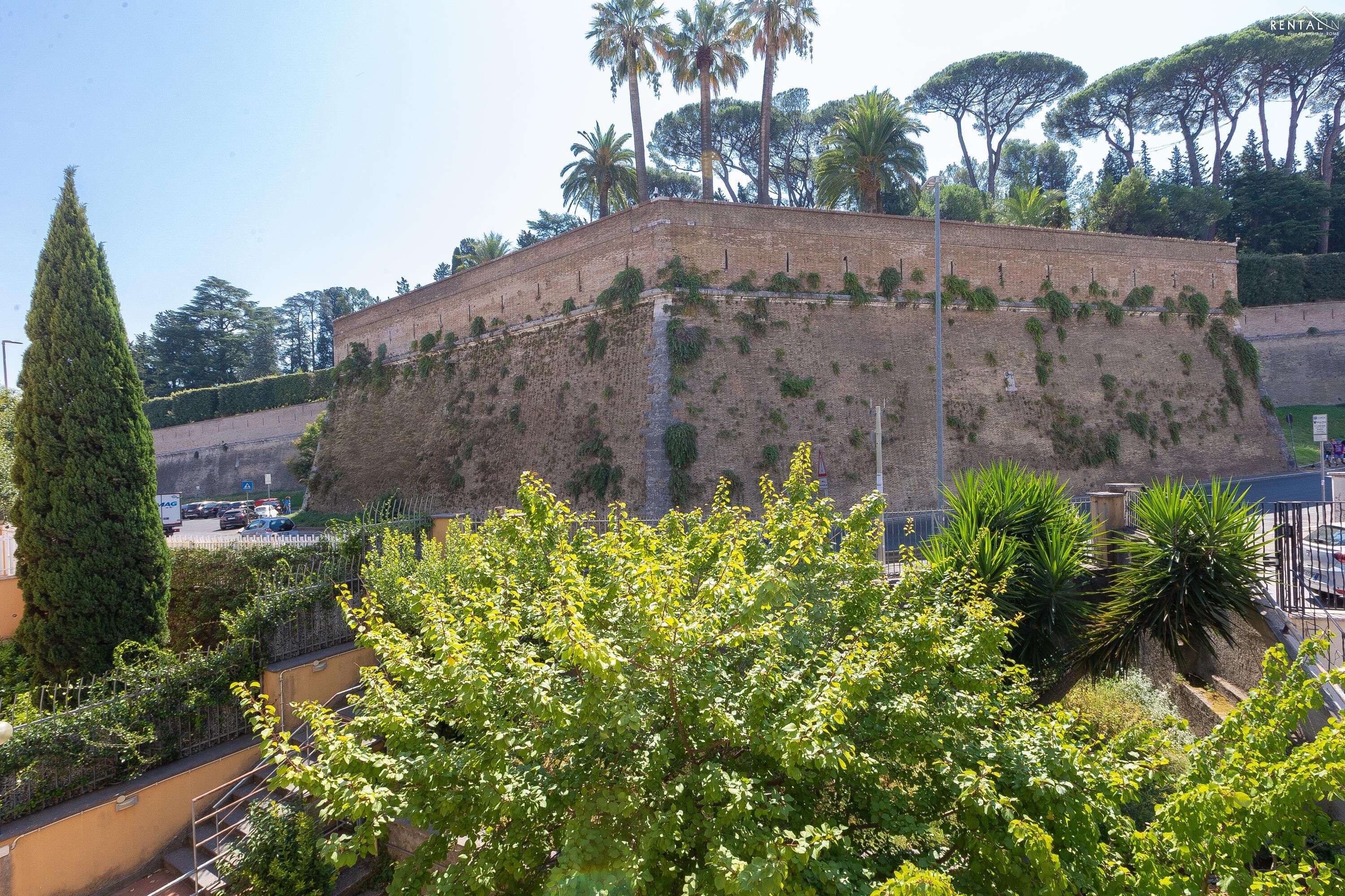 Eine historische Steinmauer mit bewuchernder Vegetation, umgeben von Bäumen und einem klaren Himmel.