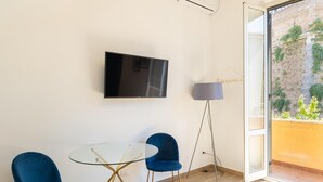 Une chambre moderne avec un téléviseur à écran plat fixé au mur, deux chaises bleues disposées autour d’une table en verre et un lampadaire.