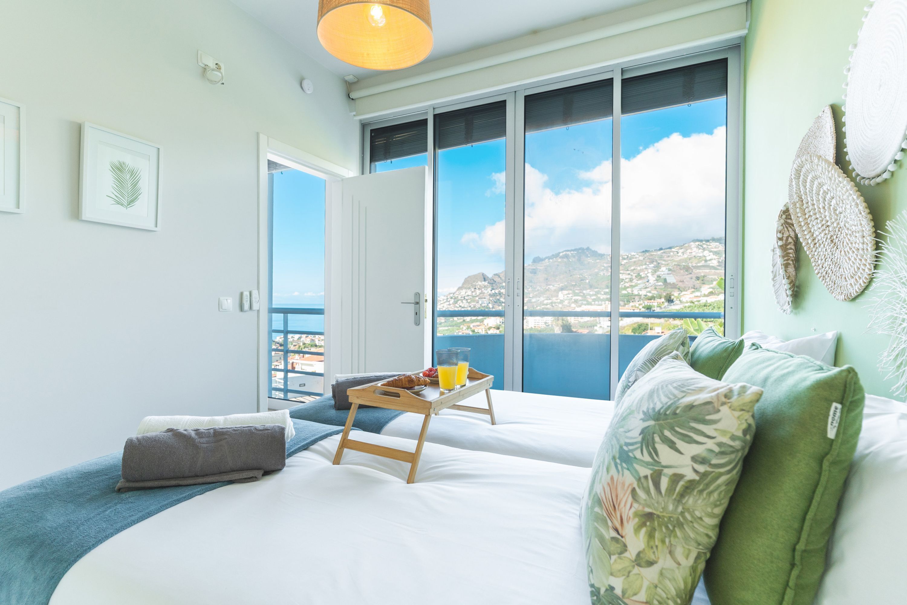 A bedroom with a large window, a bed, a bedside table with breakfast, and a view of a coastal landscape.