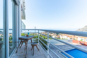 Ein Balkon mit Tisch und Stühlen, ein Pool und Blick auf eine Küstenstadt.