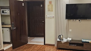 A spacious living area with a wooden floor, a dark wooden door, a wall-mounted flat-screen TV, a small table with a kettle, and a microwave in the corner. - Abha lamar for serviced apartments (Riyadh)