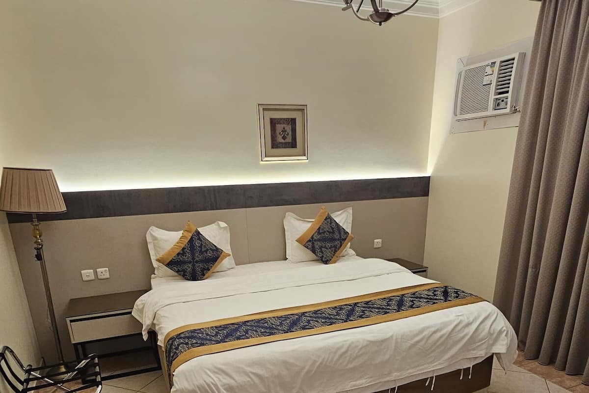 Ein Hotelzimmer mit Bett, Schreibtisch, Stuhl, Lampe, Deckenleuchte, einem Bild an der Wand und einer Klimaanlage.