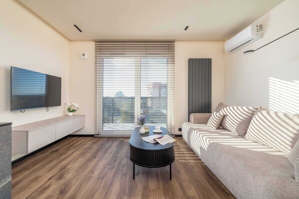 A modern living room with a flat-screen TV, a round coffee table, a sofa, and a large window with blinds.
