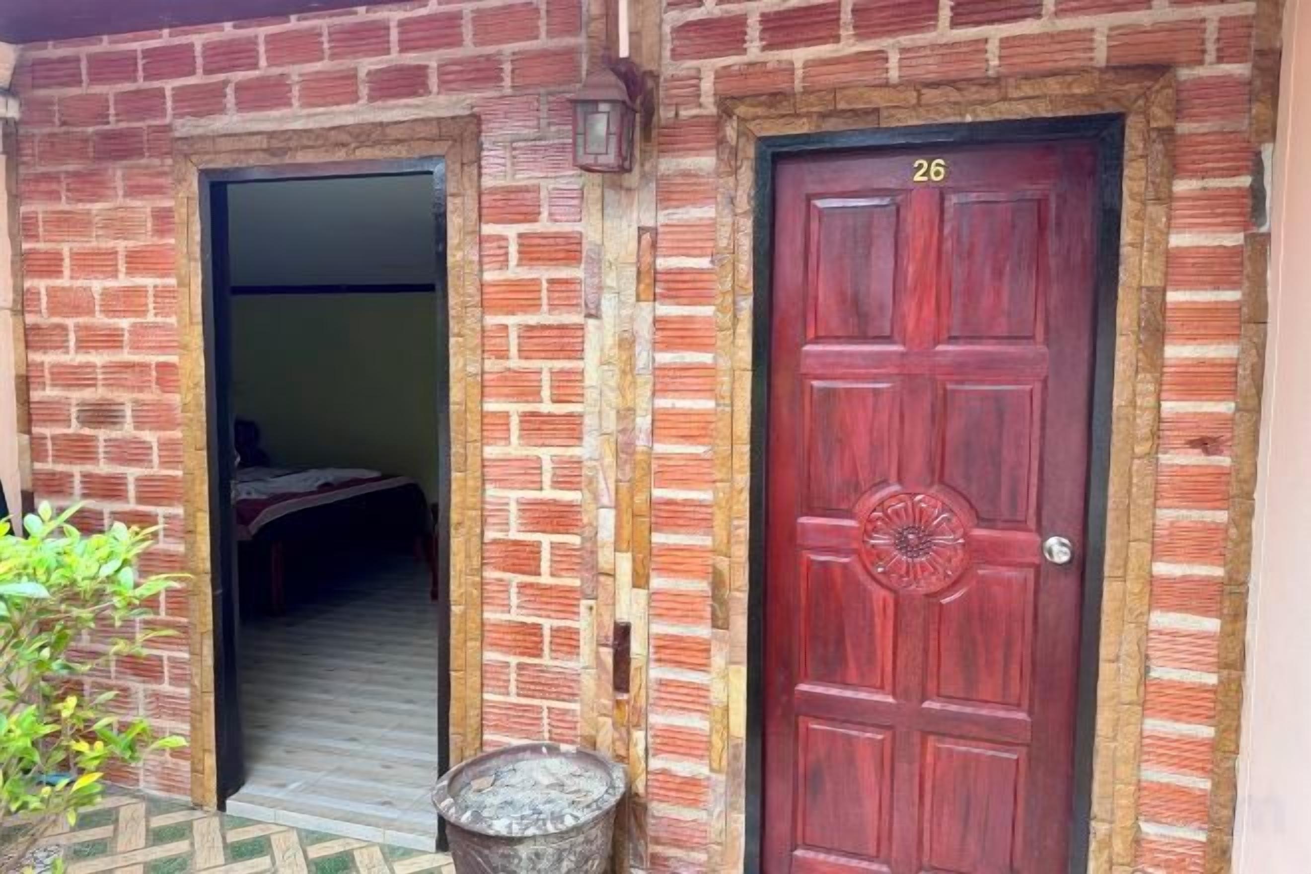 Un bâtiment en briques avec une porte rouge portant le numéro 26, un seuil donnant accès à un espace intérieur et un pot de fleurs placé à gauche.