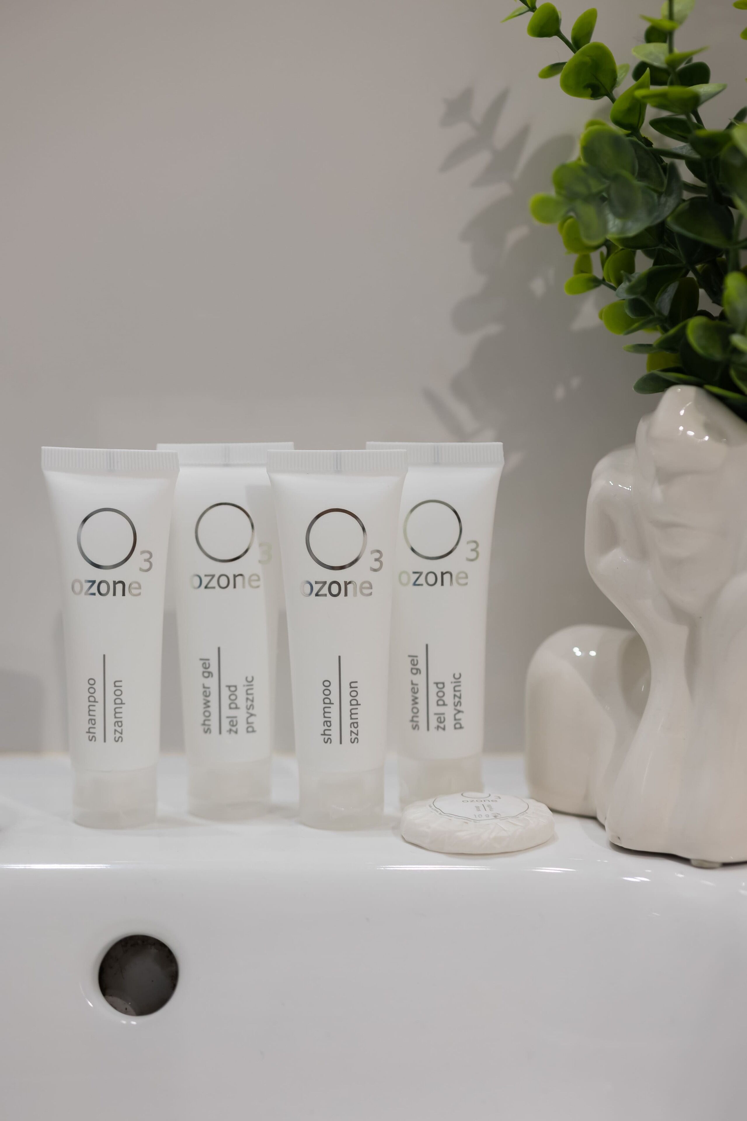 Cuatro tubos de gel de ducha y champú Ozone 3 sobre una superficie blanca, con una escultura de cerámica blanca y una planta en maceta al fondo.
