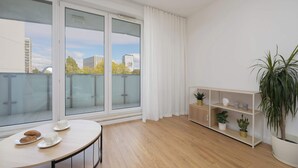 Ein modernes Apartment mit einem großen Schiebeglasfenster zum Balkon, einem runden Holztisch mit Tassen und einem Keks sowie einer weißen Regaleinheit mit Pflanzen.