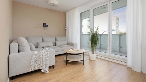 Ein modernes Wohnzimmer mit einem grauen Sofa, einem runden Couchtisch und einem großen Fenster mit Vorhängen.