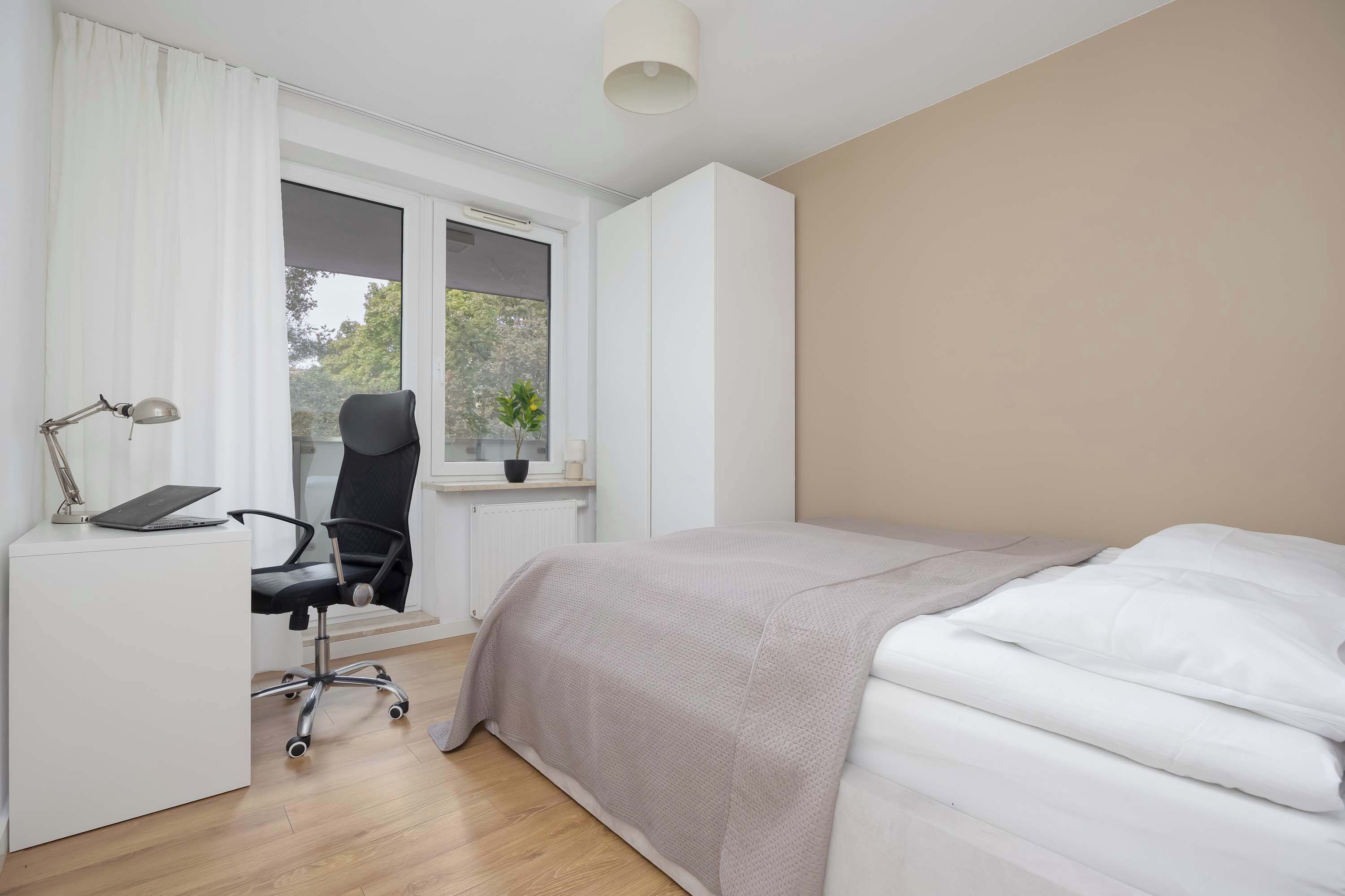 A bedroom with a bed, a desk with a chair, a window, and a view of trees outside.