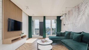A modern living room with a sectional sofa, a flat-screen TV, a small round table, and a large window offering a city view.