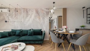 A modern living room with a green sofa, a dining table with chairs, and a kitchen area visible in the background.