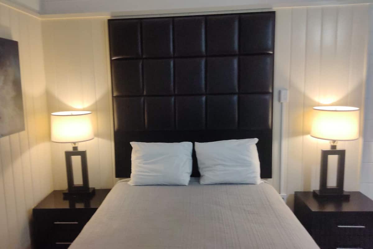 A neatly made bed with white linens and two pillows, flanked by two bedside tables with lamps, in a room with a dark headboard and a framed picture on the wall.