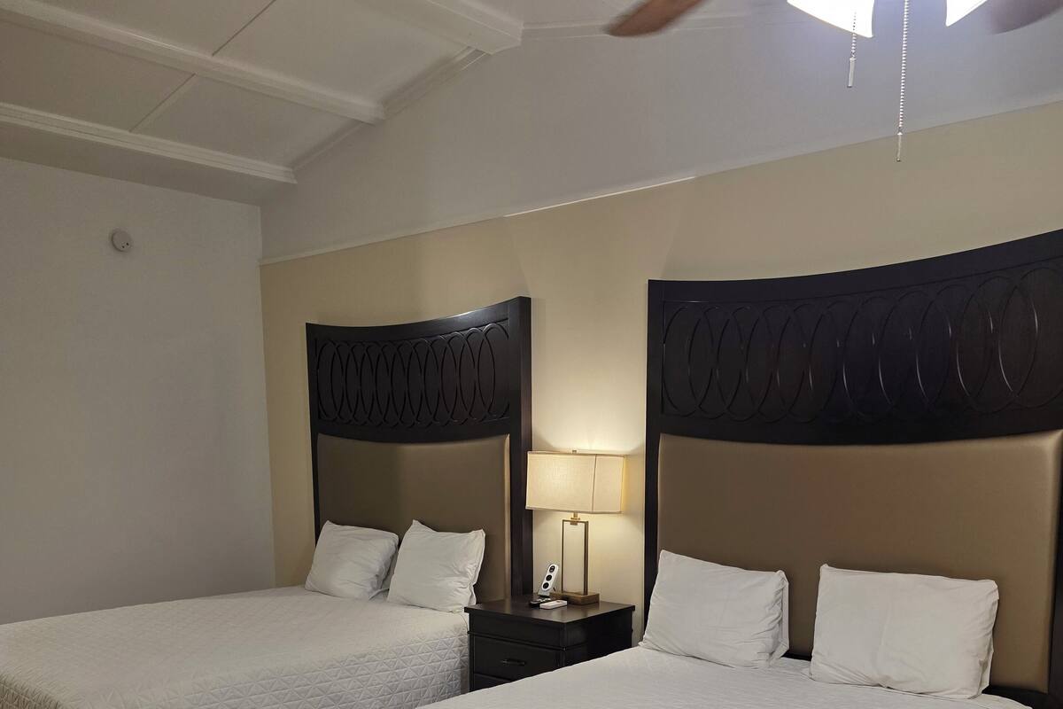 A hotel room with two beds, each with a headboard and white bedding. There is a nightstand with a lamp between the beds. The room has wooden flooring and a ceiling fan.