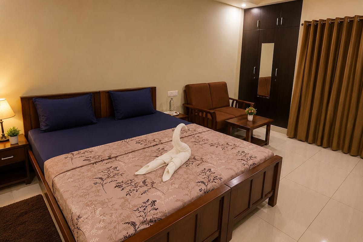 A hotel room with a bed, a chair, a small table, a nightstand, a lamp, a potted plant, a mirror, and a curtain.