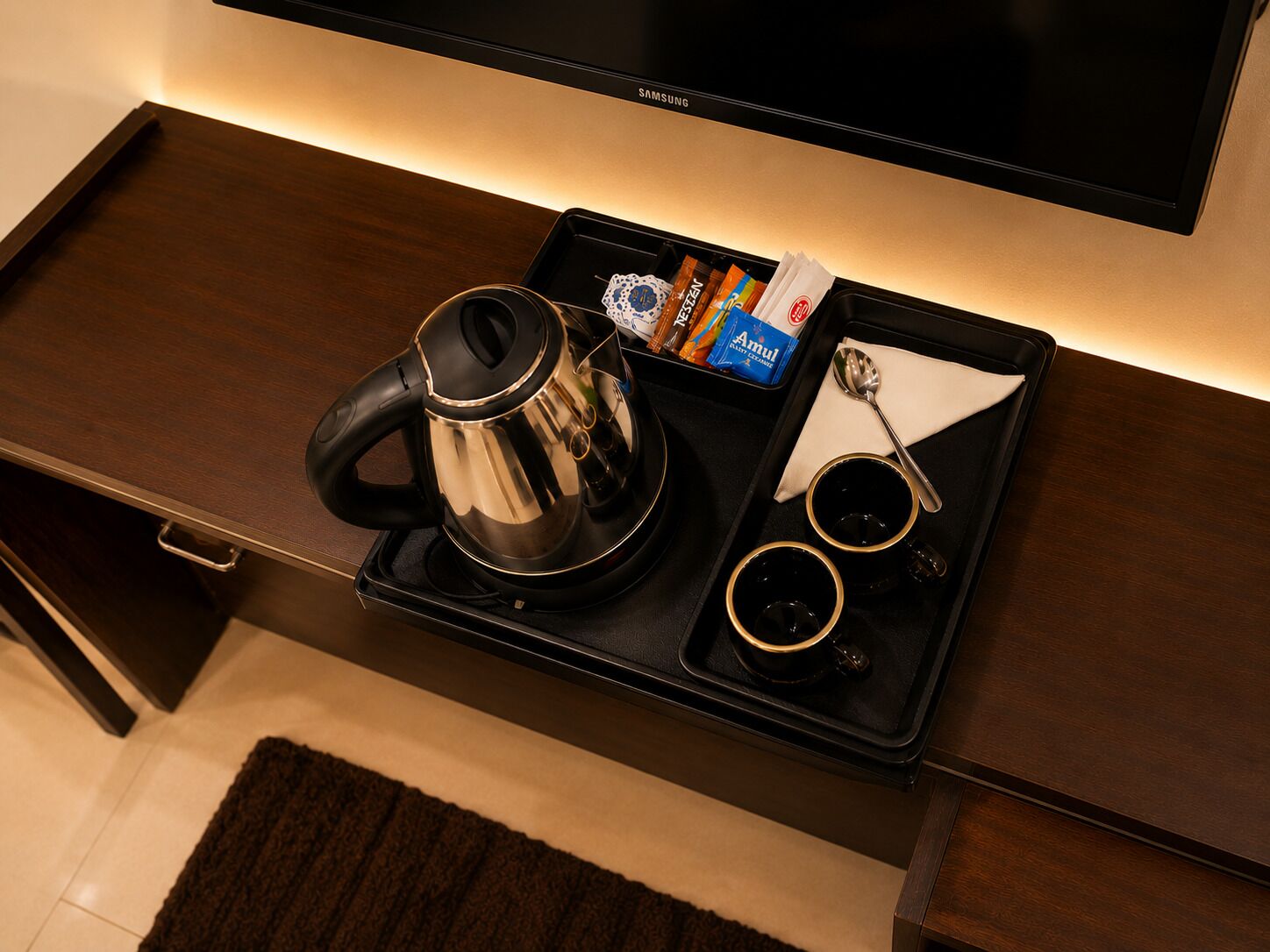 Une chambre d’hôtel équipée d’un téléviseur à écran plat fixé au mur, d’un plateau avec une bouilloire, deux tasses, une cuillère et quelques en-cas emballés.