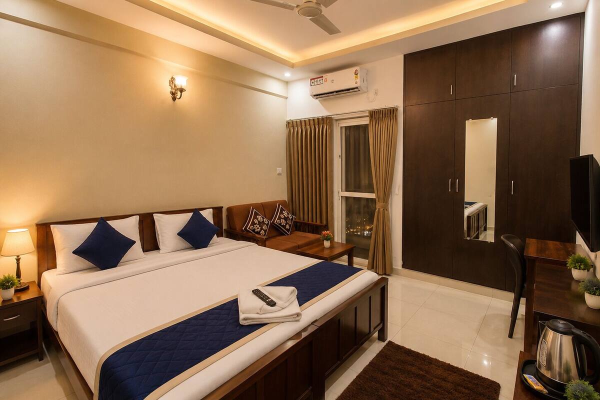 A hotel room with a bed, bedside table, lamp, air conditioning unit, wardrobe, and a view of the city.