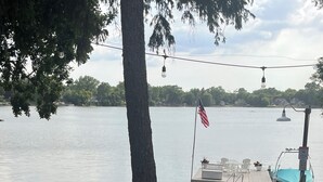A lakeside deck with potted red flowers, a dock with a boat, and an American flag.