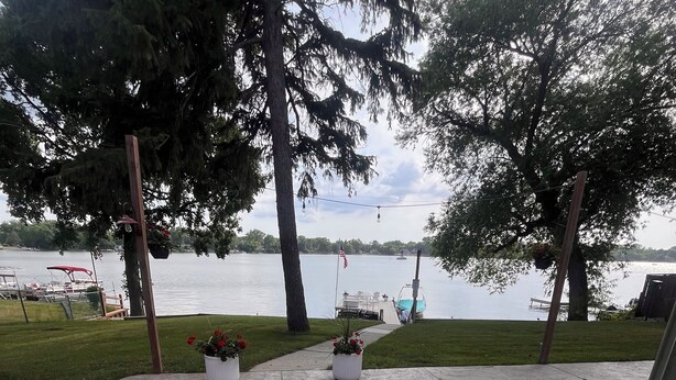 A patio with two white chairs and red cushions, overlooking a lake with boats and a dock.