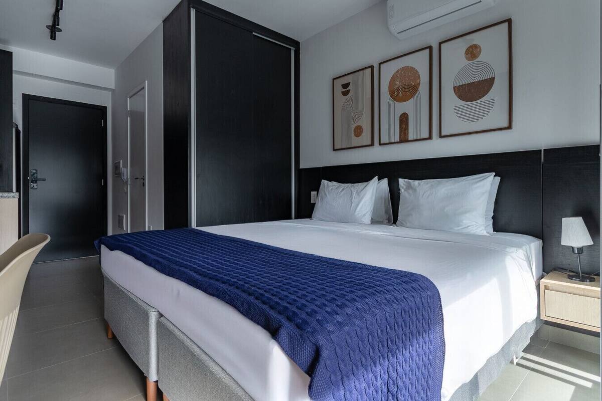Une chambre d’hôtel moderne avec un grand lit, une armoire, un bureau et une chaise. Trois œuvres d’art encadrées ornent le mur.