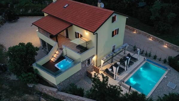 Una casa de dos plantas con piscina, mobiliario de exterior y techo de tejas rojas.