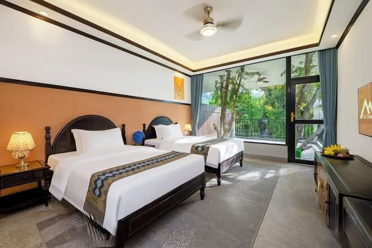 A hotel room with two beds, a desk, a chair, a lamp, and a large window with a view of greenery.