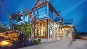 A modern house with large windows and a well-lit interior, surrounded by tropical plants and a decorative rock feature. - Fu Er Man Hotel (Luang Prabang)