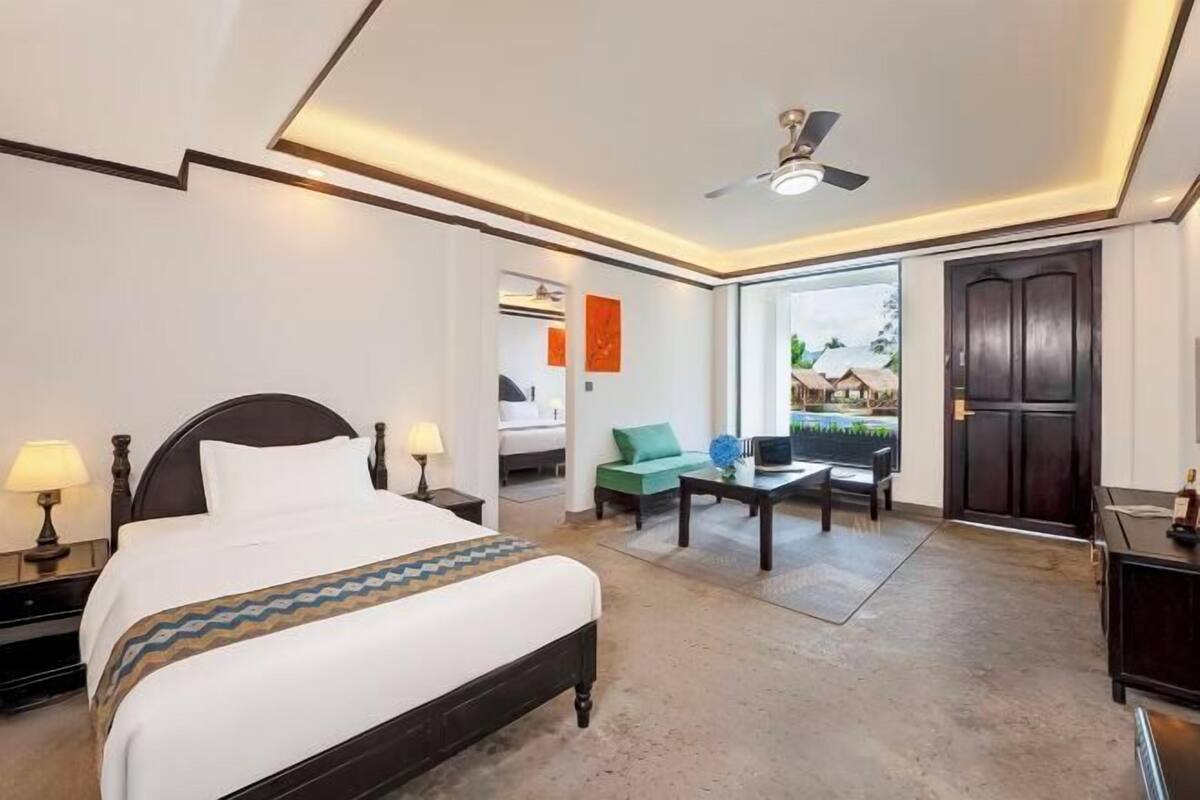 A hotel room with a bed, bedside tables, a desk, a chair, a ceiling fan, and a view of the outdoors.