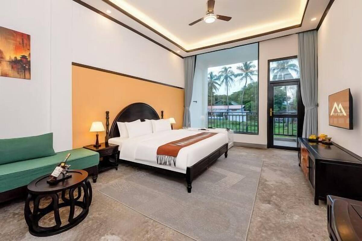 A hotel room with a bed, a green sofa, a small round table, a ceiling fan, and a large window with a view of palm trees.