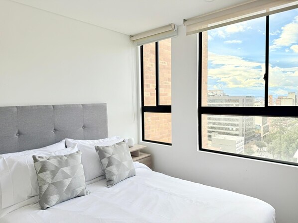 A modern hotel room with a large window, a grey upholstered headboard, and a neatly made bed with white linens and patterned pillows.