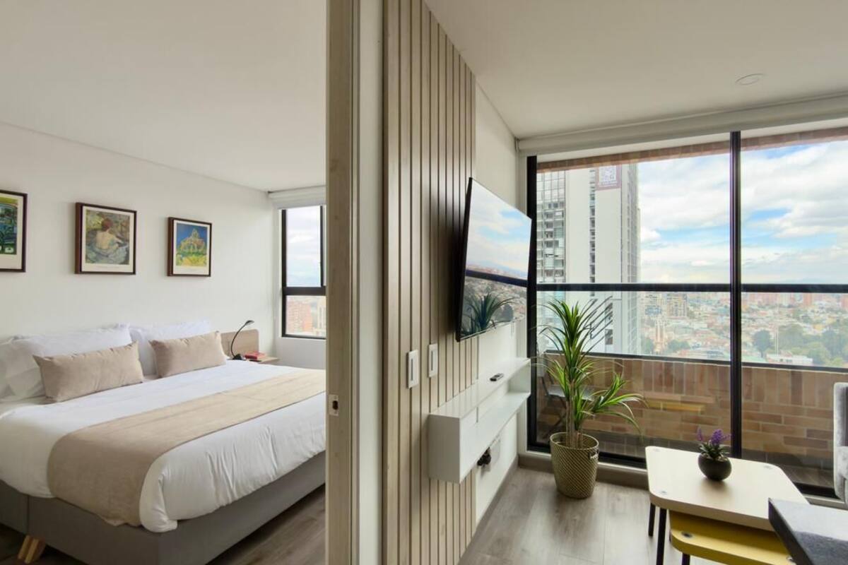 Une chambre d’hôtel moderne avec un grand lit, un téléviseur fixé au mur, un balcon offrant une vue sur la ville et une petite table agrémentée d’une plante.