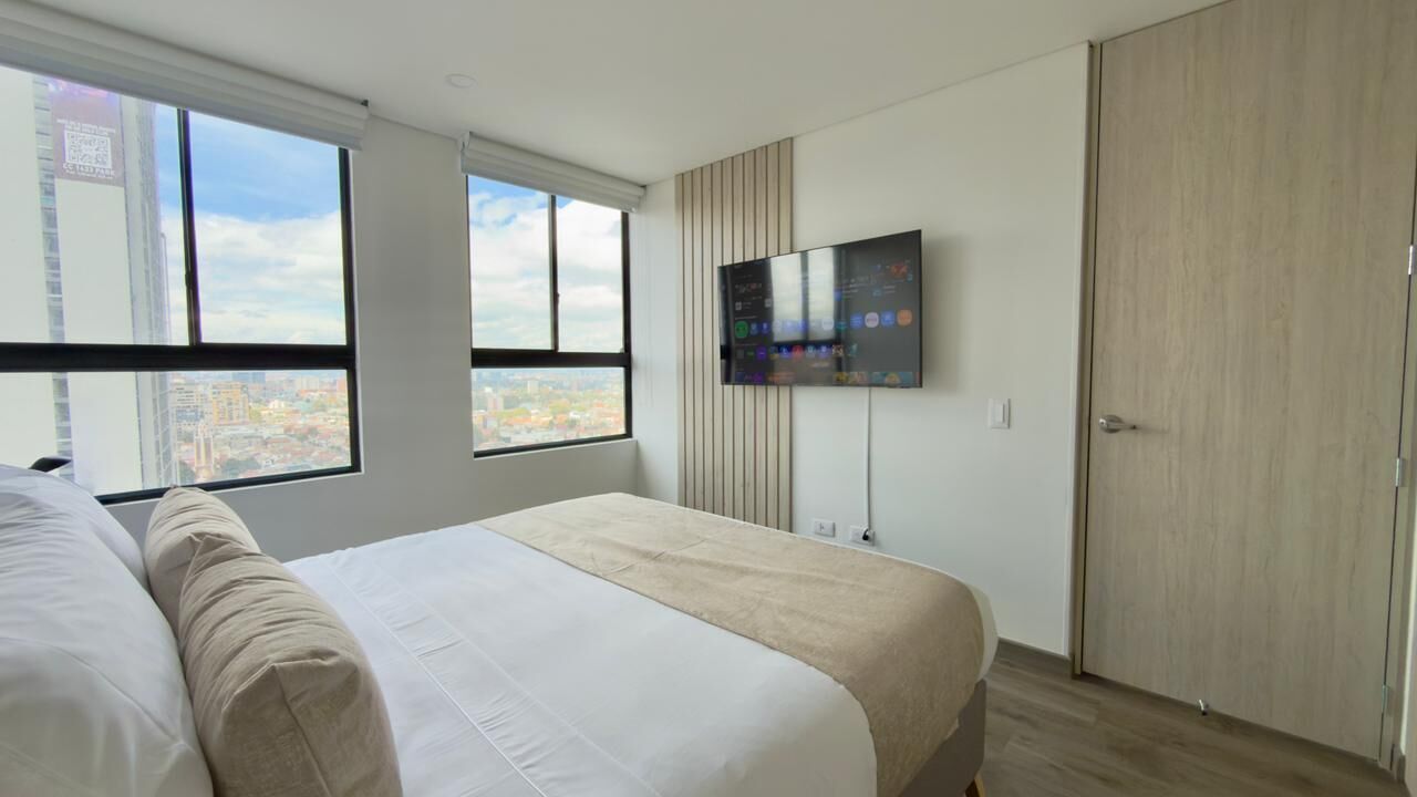 A modern bedroom with a large bed, a TV mounted on the wall, and a city view through the windows.