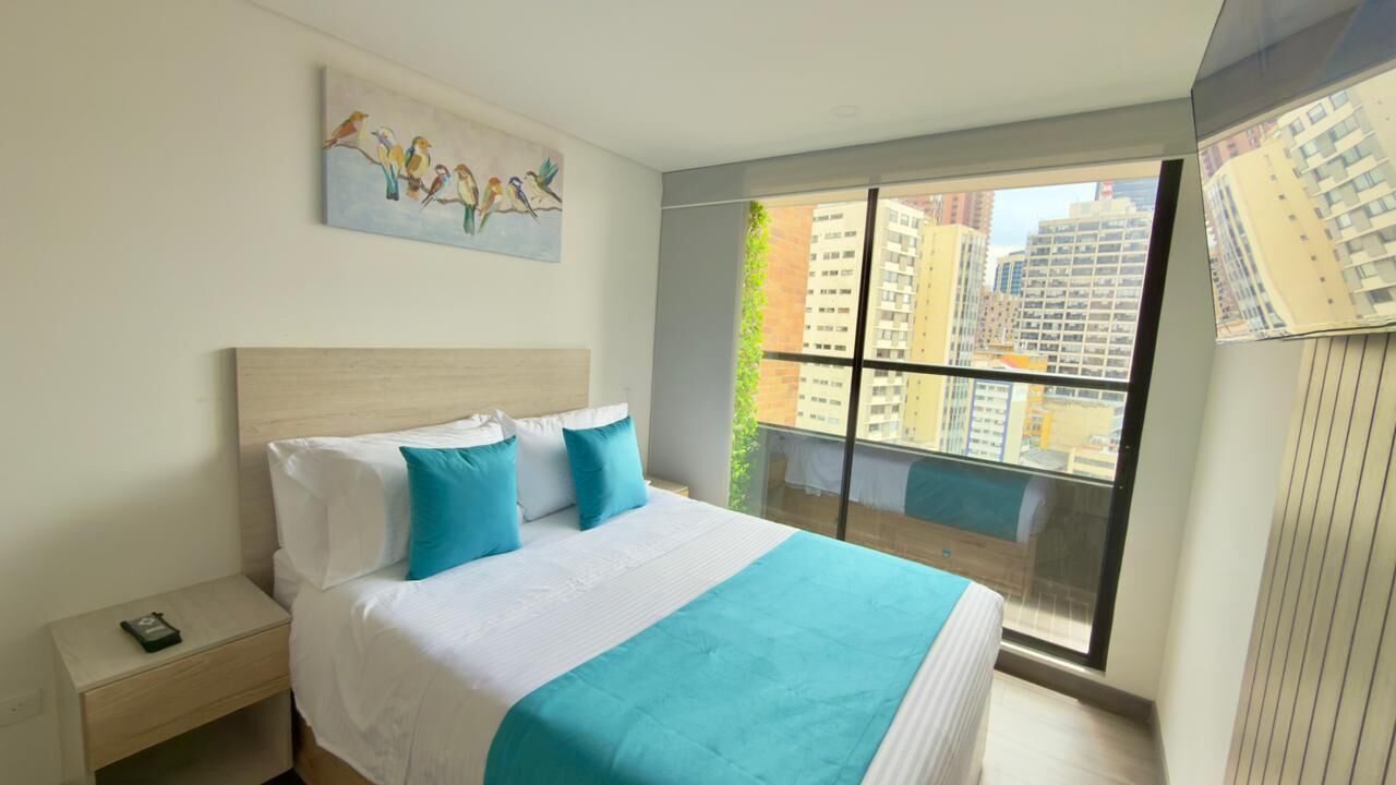 Une chambre d’hôtel moderne avec un grand lit, une table de chevet, un tableau représentant des oiseaux et une vue sur la ville par une grande fenêtre.