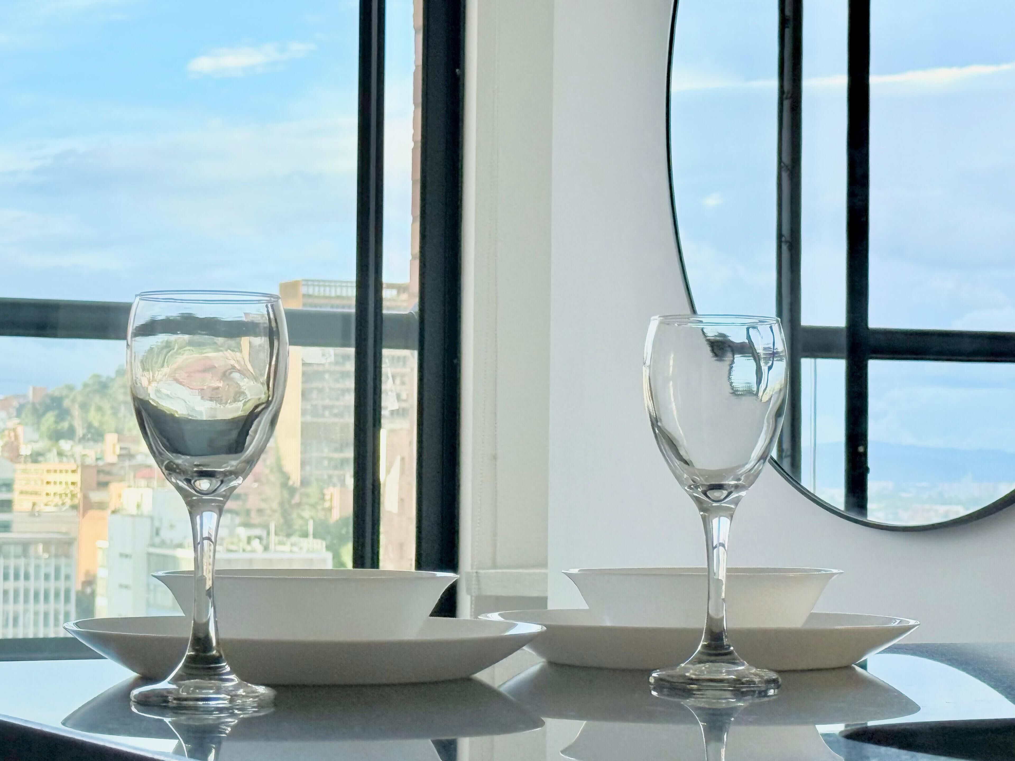 Une table avec deux verres à vin et deux bols blancs, placée devant une fenêtre donnant sur un paysage urbain.