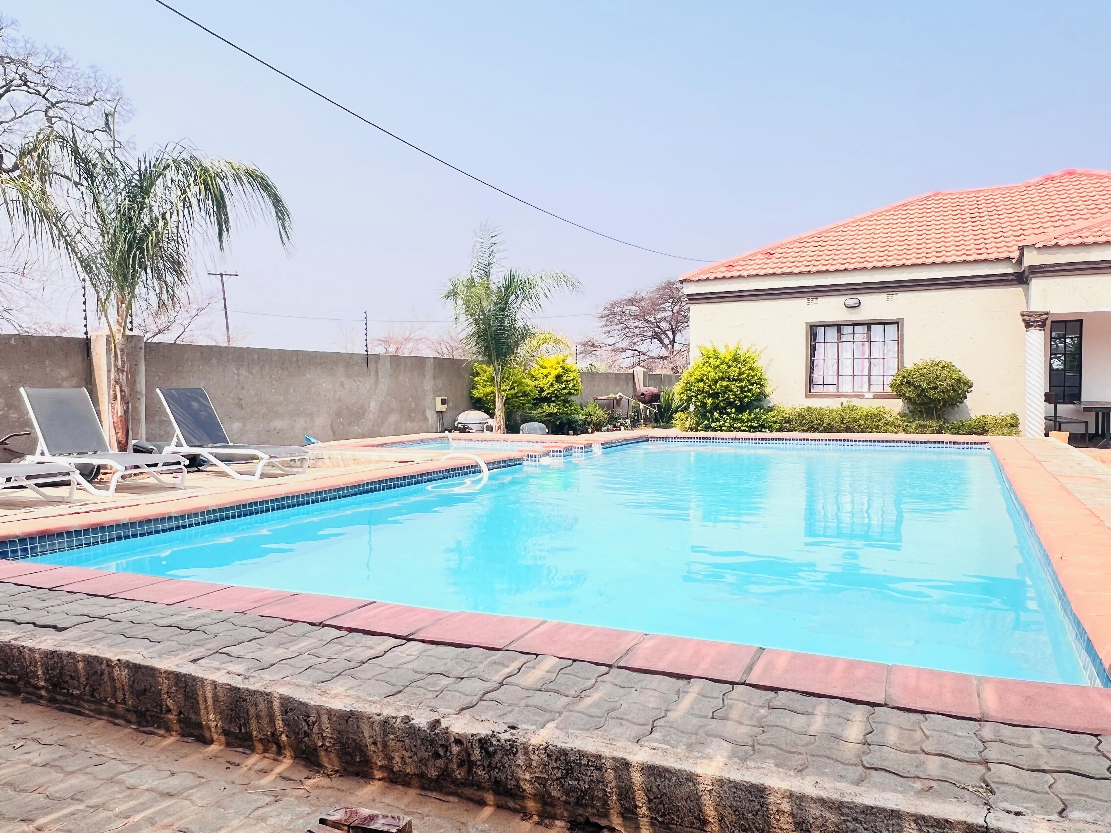 Une piscine rectangulaire dont le bord est recouvert de carrelage, entourée d’une terrasse dallée. On aperçoit, au fond, des chaises longues et un barbecue.