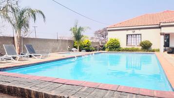 Une piscine rectangulaire dont le bord est recouvert de carrelage, entourée d’une terrasse dallée. On aperçoit, au fond, des chaises longues et un barbecue.