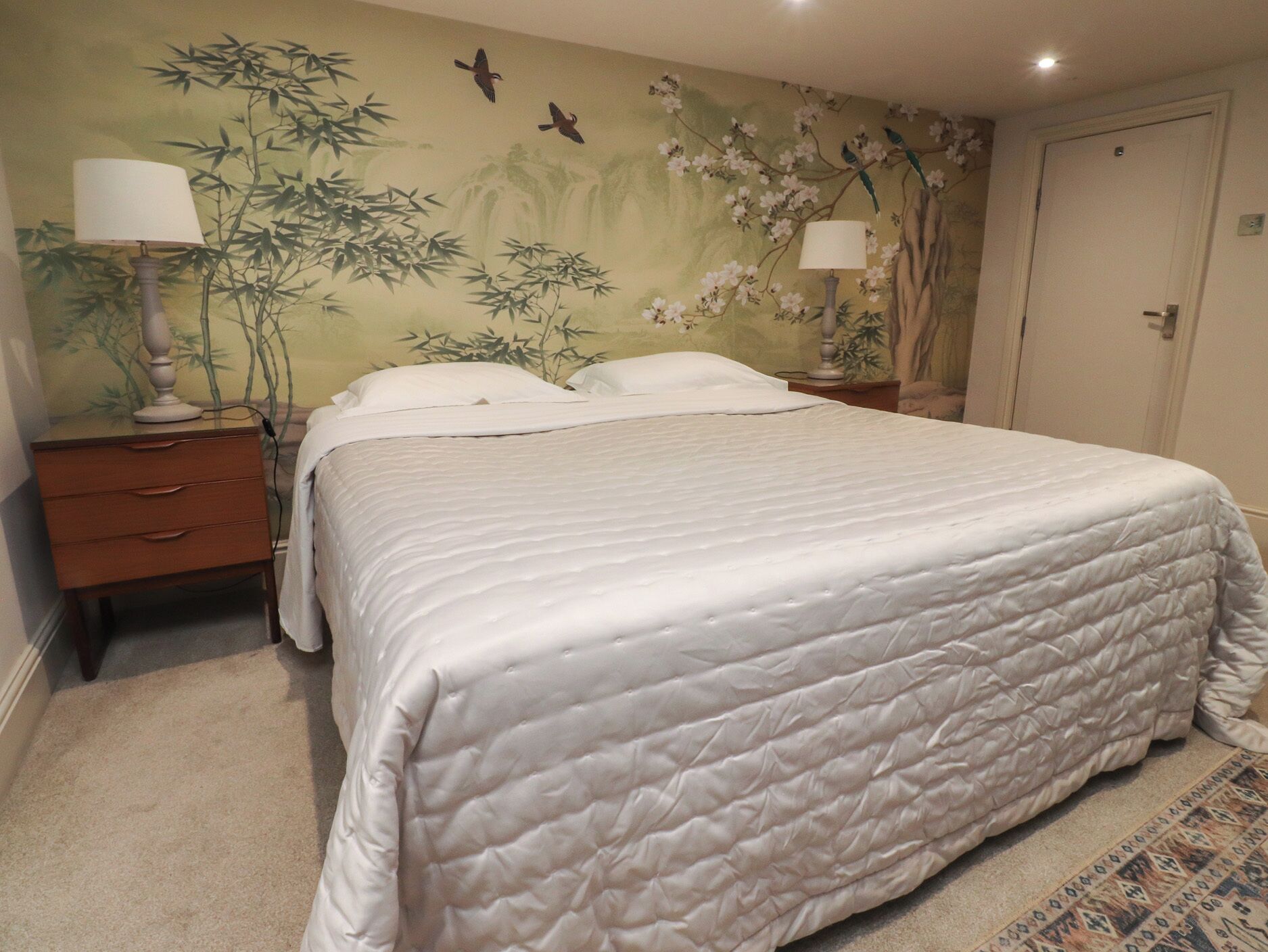 A neatly made bed with a quilted white duvet, a wooden nightstand with a lamp, and a wall with a floral and bamboo mural.