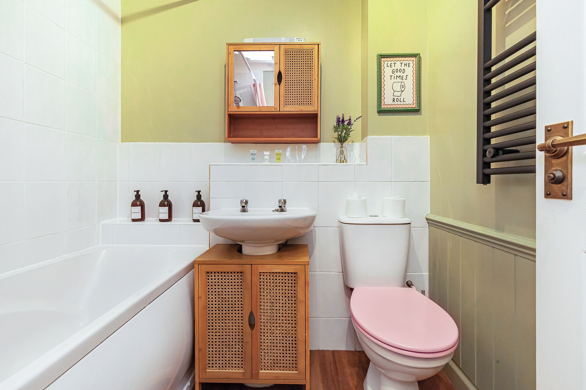 Ein Badezimmer mit einer weißen Badewanne, einem Holfach mit einer Rattanschranktür, einem weißen Waschbecken, einer Toilette mit rosa Sitz und einem gerahmten Schild an der Wand.