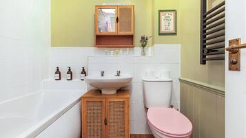 Ein Badezimmer mit einer weißen Badewanne, einem Holfach mit einer Rattanschranktür, einem weißen Waschbecken, einer Toilette mit rosa Sitz und einem gerahmten Schild an der Wand.