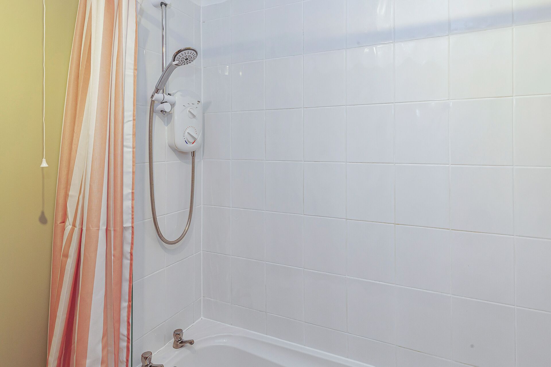 Une salle de bain avec des murs recouverts de carreaux blancs, une douche avec un pommeau de douche amovible et une baignoire.