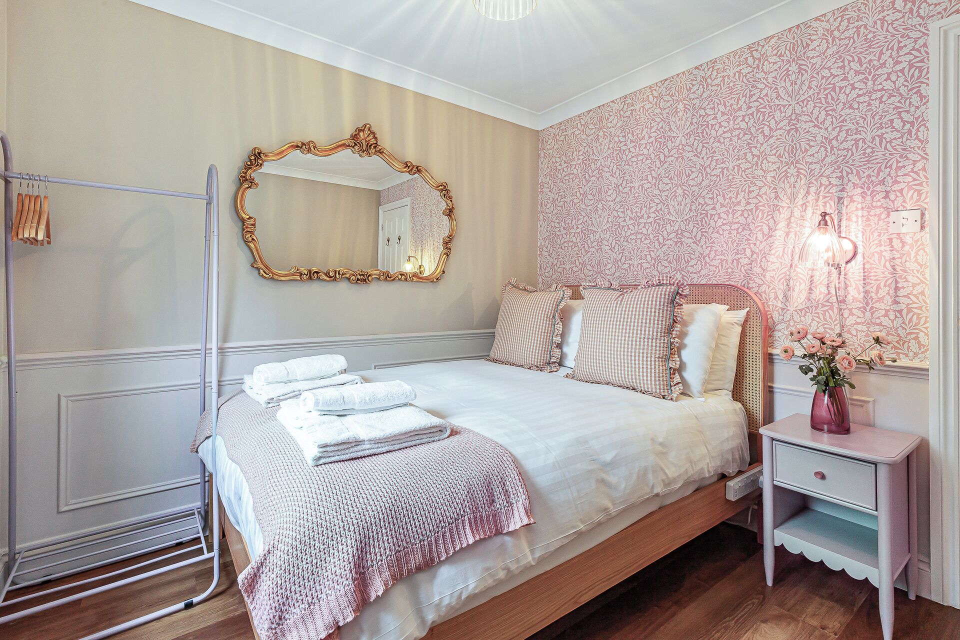 Una cama bien hecha con edredón blanco y manta de punto rosa, una mesita de noche con lámpara, y un espejo en la pared.
