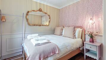 Una cama bien hecha con edredón blanco y manta de punto rosa, una mesita de noche con lámpara, y un espejo en la pared.