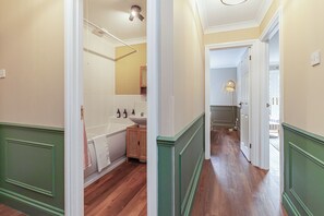Een gang met houten vloeren en groene lambrisering, die uitkomt bij een badkamer met bad en wastafel.
