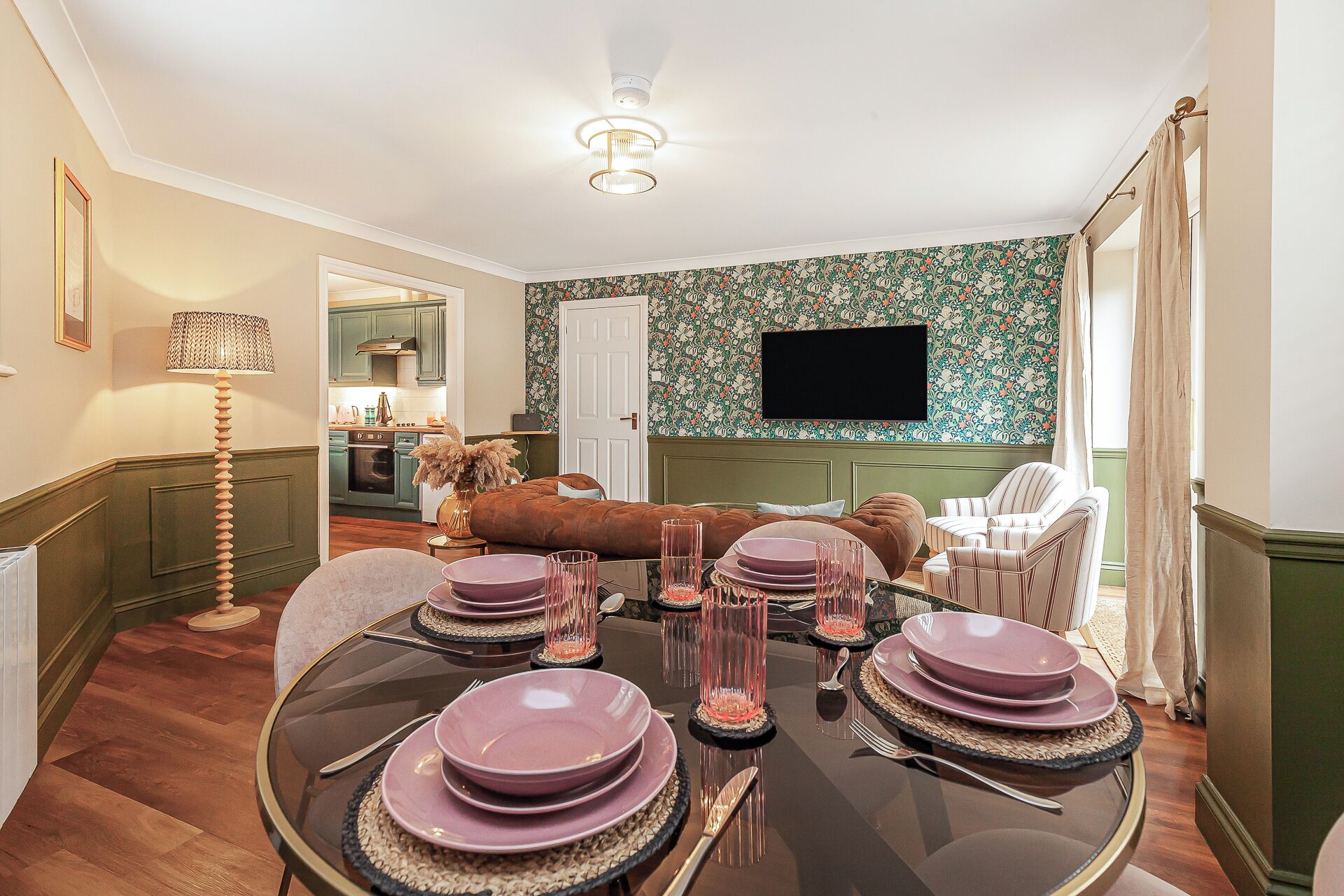 Un coin repas avec une table ronde dressée pour quatre personnes, un téléviseur fixé au mur et une cuisine visible au fond.