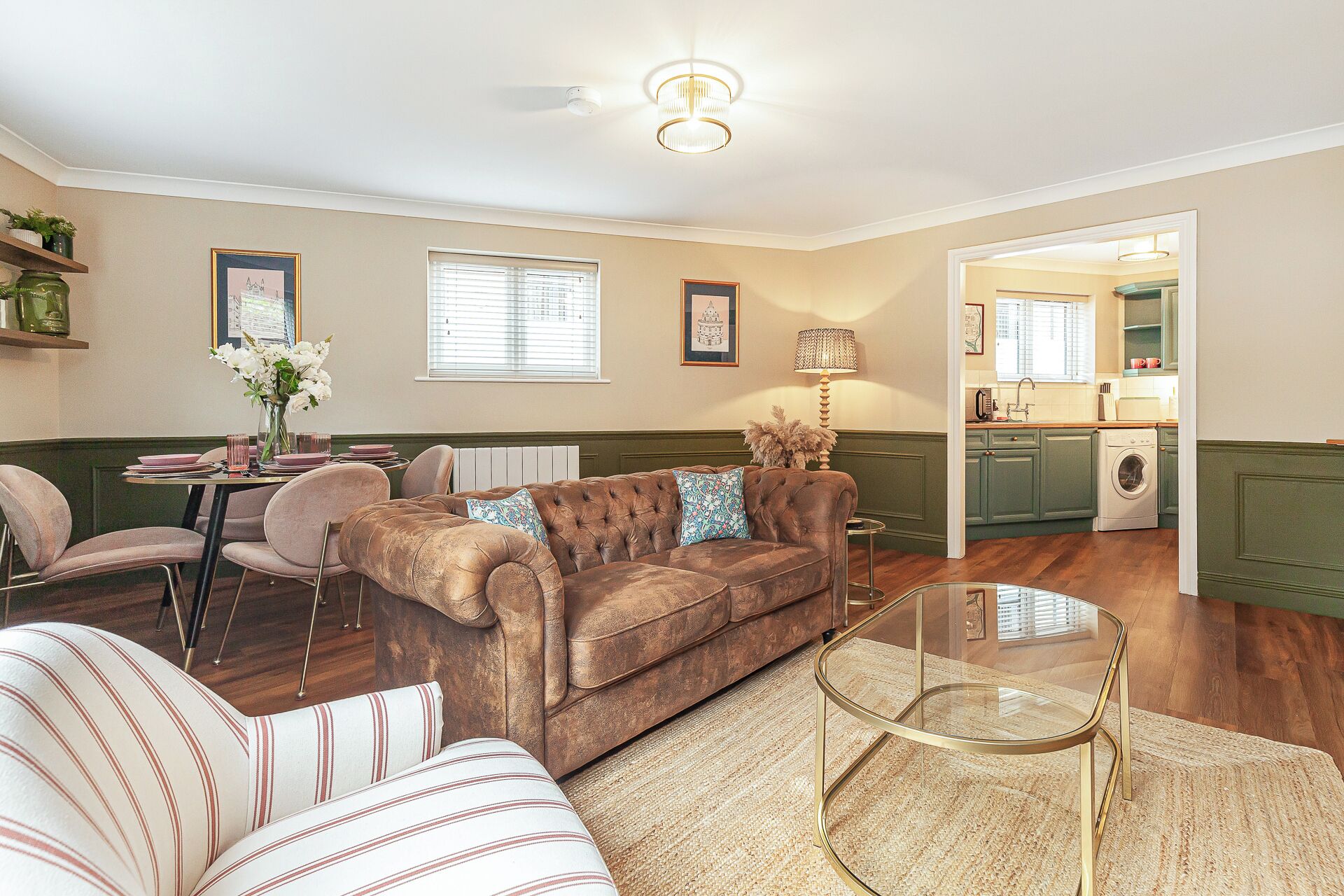 A living room with a brown sofa, striped armchair, and a glass coffee table. There is a dining area with pink chairs and a table, and a kitchen with green cabinets and a washing machine.
